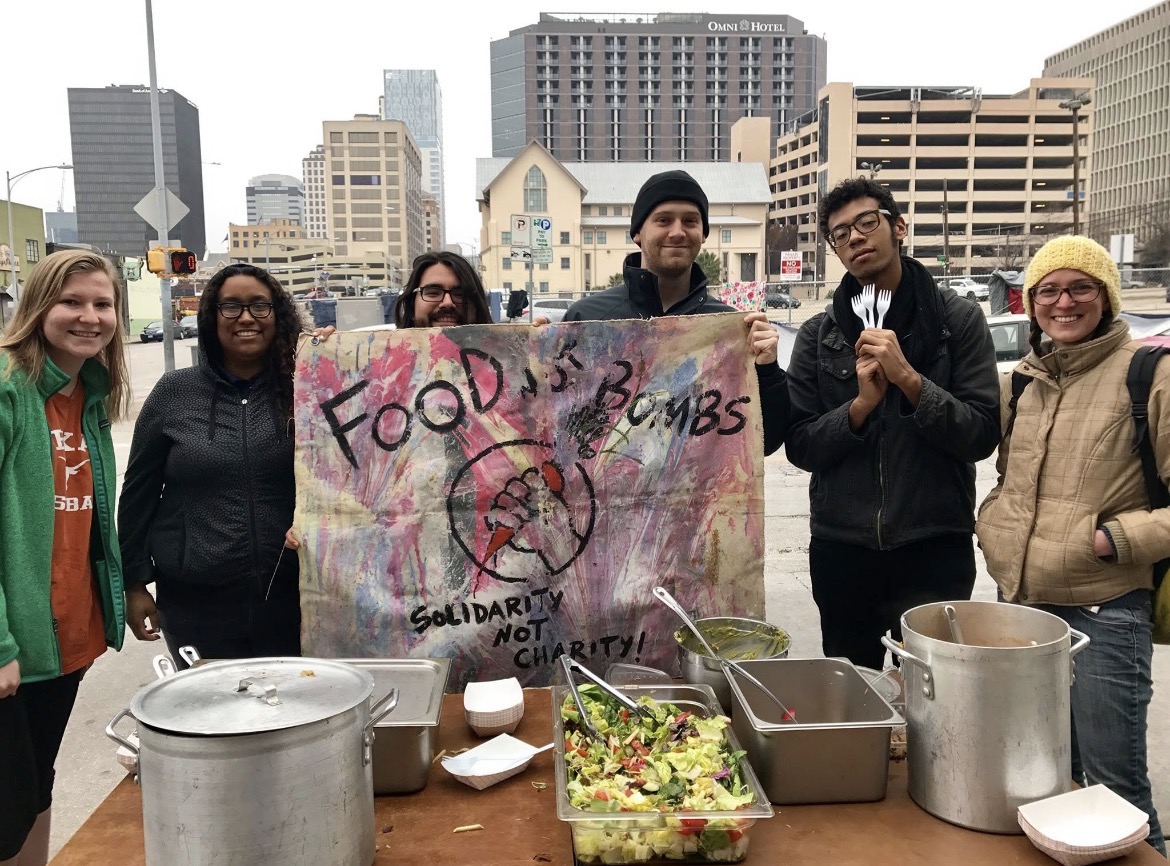 Volunteers holding a hand-painted Food Not Bombs banner behind trays of food in downtown Austin