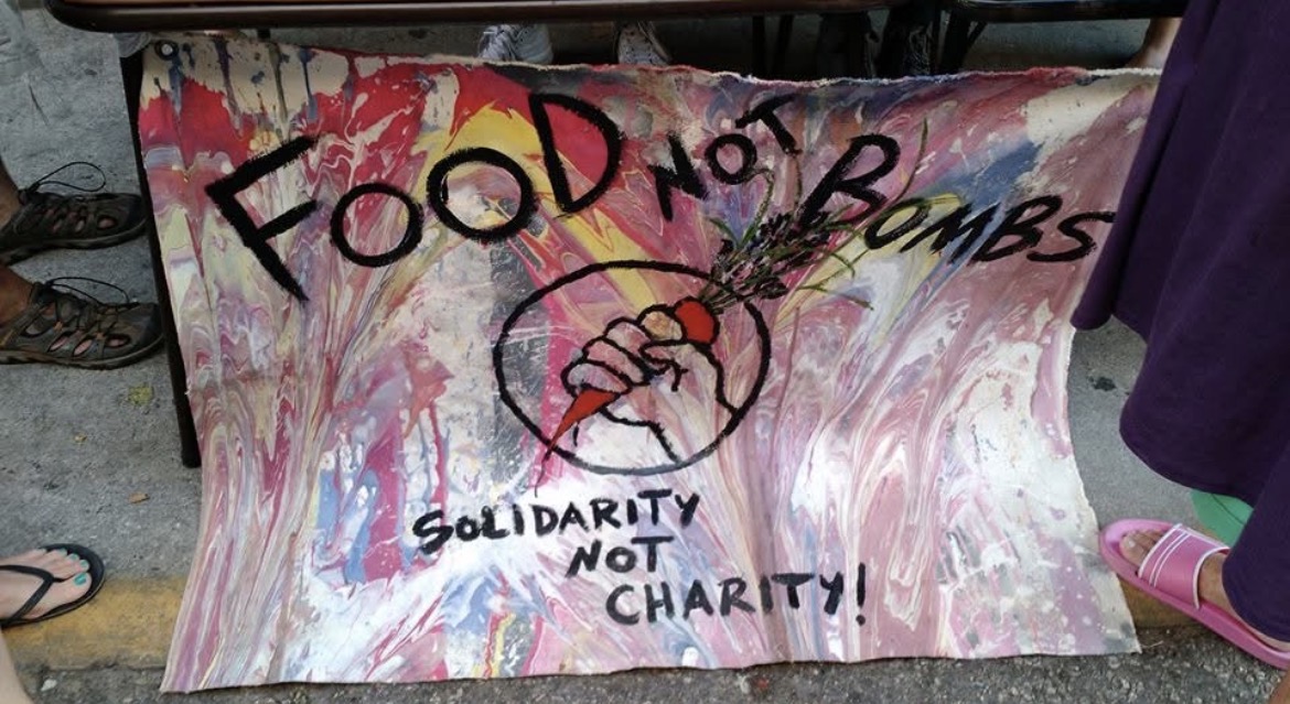 Hand-painted Food Not Bombs banner reading Solidarity Not Charity with a fist holding a carrot